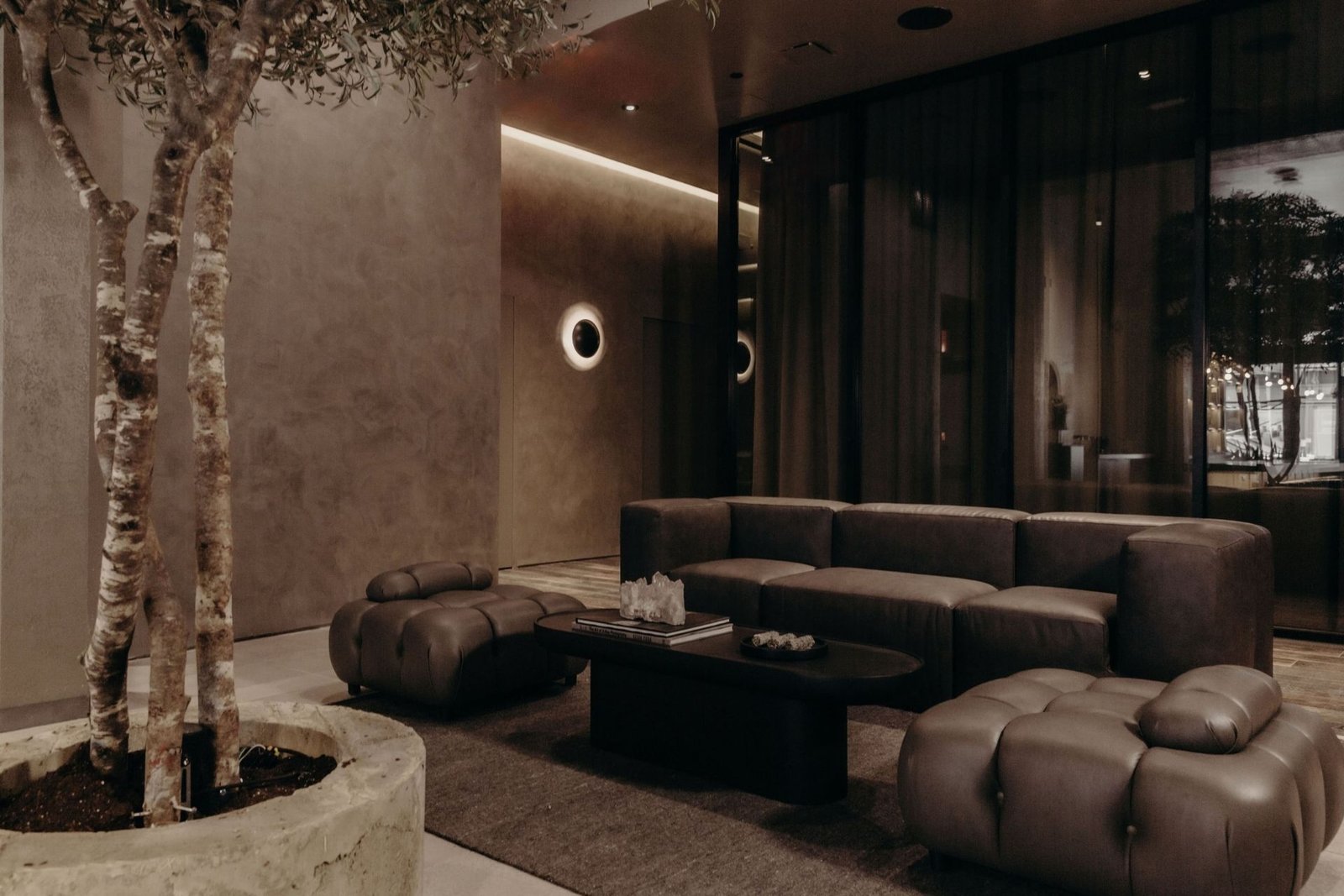 Remedy Place evening lounge with tufted leather sofa, olive tree, halo sconce and city lights beyond the glass