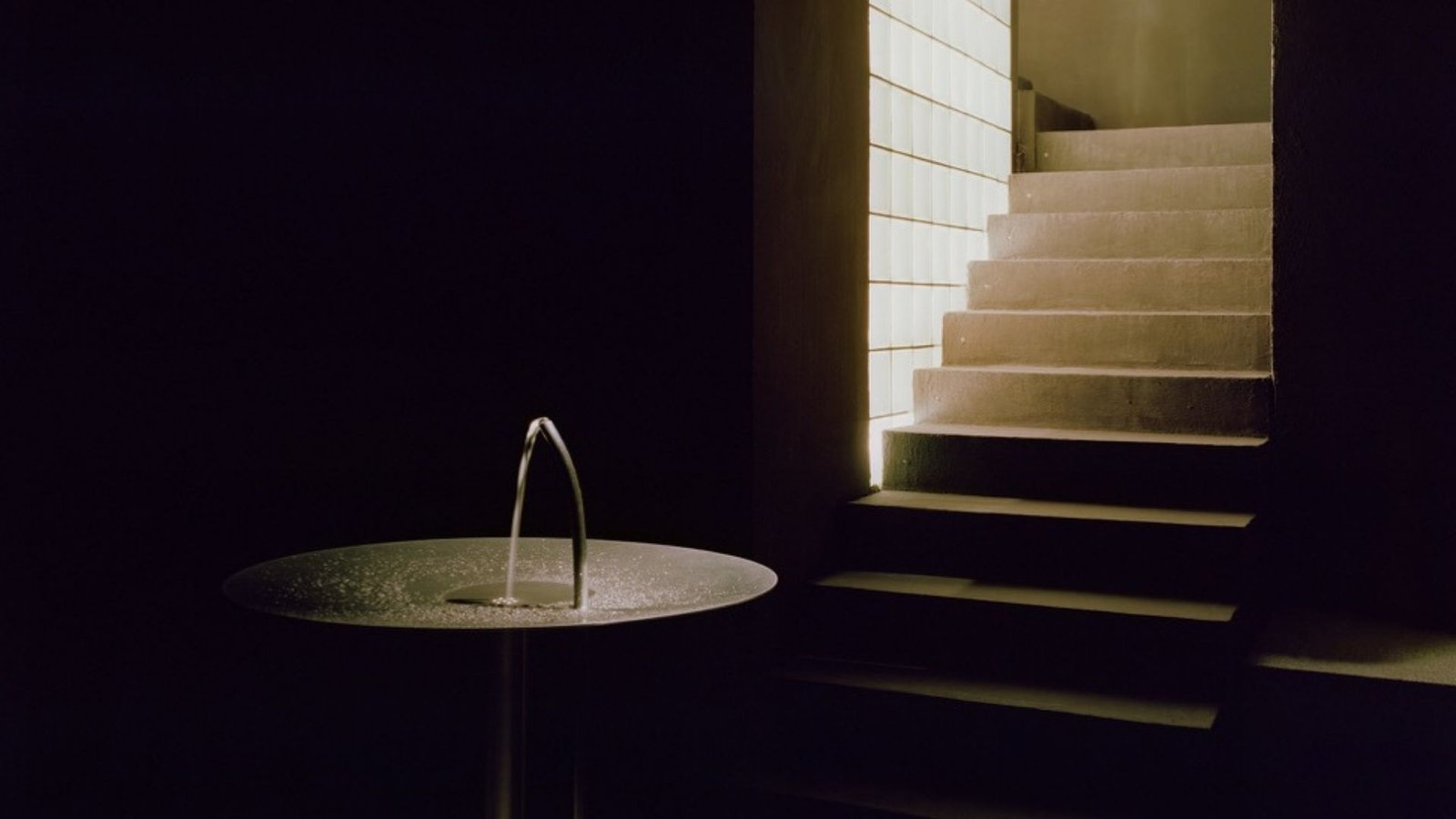 A fountain arc and an ascending staircase lit by an amber glass-block wall at Riela Madrid
