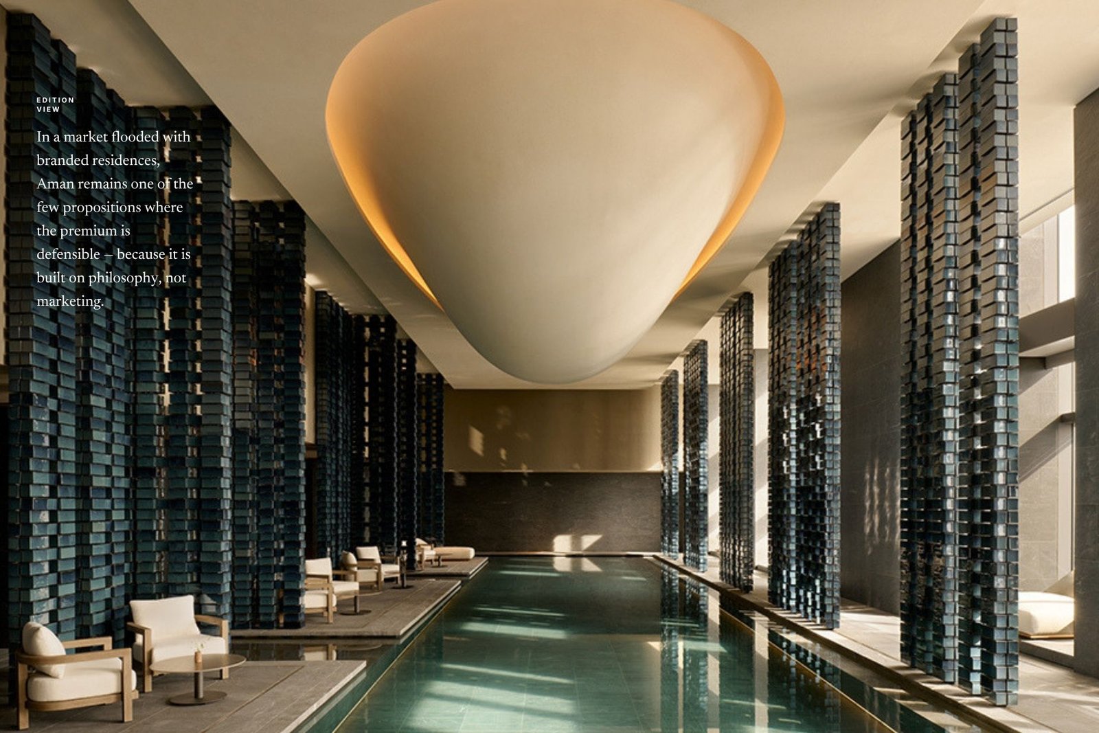 A symmetrical view down an Aman indoor lap pool with teal water and blue-glazed tiled columns beneath a glowing sculptural ceiling, with an Edition Media 'Edition View' quote on Aman's defensible premium.